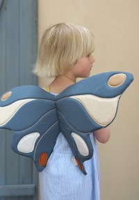 Textilní motýlí křídla v modré, krémové a hnědé barvě s vypolstrovanou texturou. Obsahují detaily a ozdoby. Nošena na zádech dítěte přes pruhované šaty.