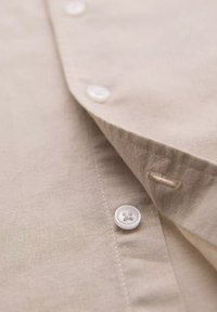 Beige linnen shirt met een opgevouwen kraag en witte knopen. Zichtbare stikdetails en een gladde textuur. De knoopsluiting is gedeeltelijk open.