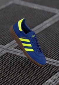 Modré semišové tenisky s třemi fluorescenčně žlutými pruhy, hnědou gumovou podrážkou a polstrovaným límcem. Obsahují klasické branding adidas a minimalizovanou šněrování.
