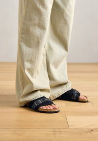 Personne portant un pantalon beige clair ample et des sandales noires à semelle en liège, se tenant sur un parquet en bois clair.