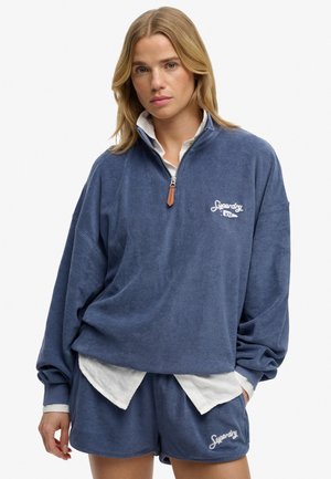 Kobieta ubrana w luźną granatową bluzę z zamkiem do ćwiartki oraz pasujące szorty z białą koszulą pod spodem, stojąca na jednolitym tle.