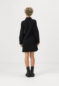 Crna traper haljina s dugim rukavima i ovratnikom, koja ima pripijeni kroj i dizajn do koljena. Nosila se s crnim gležnjačama.