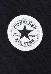 Svart tyg med en vit rund lapp som har en stjärna. Lappen inkluderar texten "CONVERSE" och "ALL STAR" i ett fetstiligt teckensnitt.