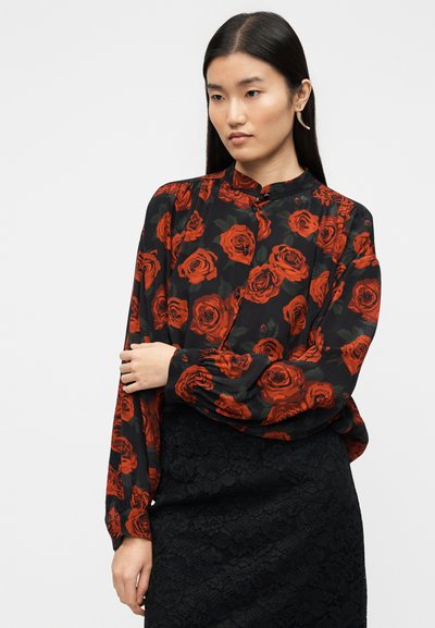 Blouse noire à motif de roses rouges vives, manches longues, col haut et détail à boutons, assortie à une jupe en dentelle noire texturée.
