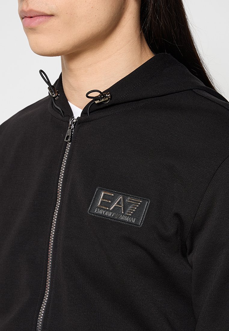 Schwarze Kapuzenjacke mit Reißverschluss und strukturiertem Patch-Logo "EA7 Emporio Armani." Verfügt über verstellbare Kordeln an der Kapuze und eine glatte Stoffoberfläche.
