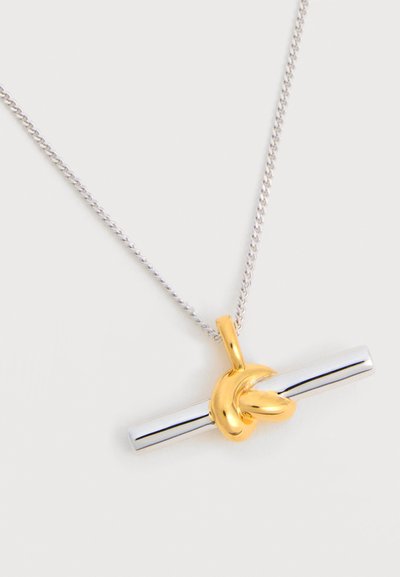 Ασημένιο κολιέ με αλυσίδα και οριζόντιο ασημένιο μενταγιόν σε σχήμα μπάρας, τυλιγμένο με λεπτομέρεια χρυσής κόμπας, σε λευκό φόντο.