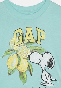 Cilvēka kora krāsas kokvilnas T-krekls ar Snoopy grafiku blakus citroniem un vārdu "GAP" treknrakstā dzeltenā krāsā.