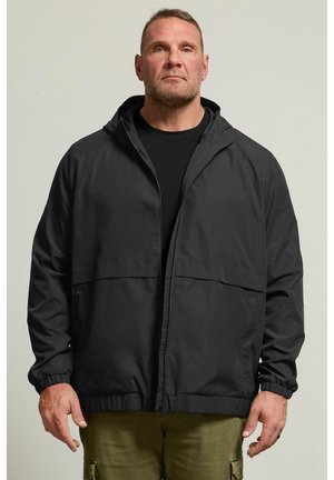 Mand iført en sort hættetrøje trukket halvt op, sort skjorte og olivengrønne bukser mod en ensfarvet lys baggrund.