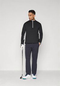 Fekete, negyedzáras pulóver szürke díszítésekkel, tengerészkék nadrág, fehér golfcipő kék részletekkel, golfütőt tartva, semleges háttér előtt állva.