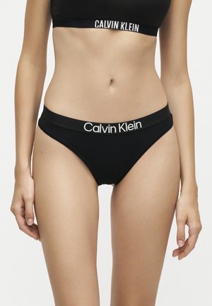 Γυναίκα που φοράει μαύρο σετ εσωρούχων Calvin Klein, παρουσιάζοντας την κοιλιά και το πάνω μέρος των μηρών σε απλό φωτεινό φόντο.