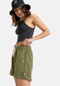 Gröna bomullsshorts med rullad fåll, sidfickor och knappdetaljer, ihop med en mörköm ribbad crop top och en svart bucket-hatt.