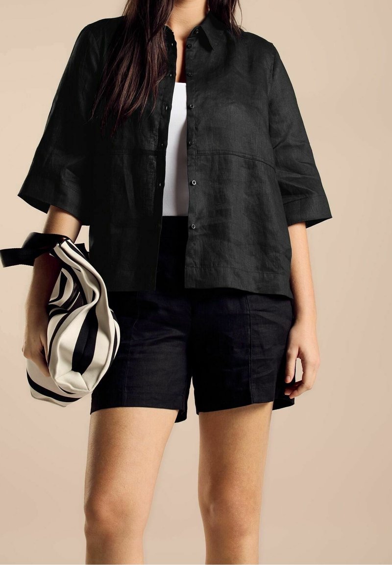 Chemise noire en lin à boutons avec manches courtes, associée à un short noir. Le modèle tient un sac cabas rayé noir et blanc.