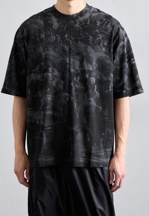 Mann trägt schwarzes übergroßes T-Shirt mit dunklem, abstraktem Menschenmuster und lockerer schwarzer Hose vor einem einfarbigen hellgrauen Hintergrund.