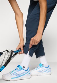 Balti sporta apavi ar ziliem akcentiem un sietam paneļiem, valkāti kopā ar baltam zeķēm. Cilvēks pielāgo piegrieztas tumši zilas bikses, turētās tenisa raketi.