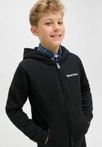 Črna zip-up kapuca iz mehkega materiala, z sprednjim žepom, rebrastimi manšetami in izvezenim napisom "Original Studio" na prsih.
