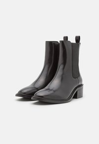 Botas de tobillo de cuero negro con acabado brillante, paneles laterales elásticos, punta afilada y tacones cuadrados. Textura suave y detalles de diseño mínimos.