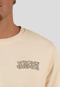 Felpa beige con testo ricamato nero "WRONG FRIENDS" indossata da una persona, che mostra il collo e parte del viso e della spalla.