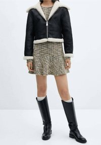 Veste en shearling noire avec fermeture éclair à l'avant ; robe mini à carreaux ; bottes noires jusqu'aux genoux ; tissu texturé ; jupe de style skater.