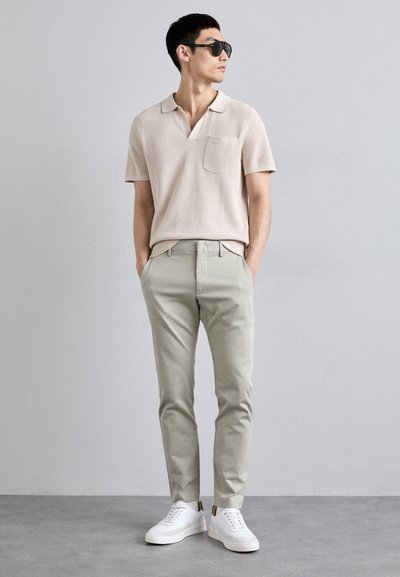 Ljus beige texturerad polotröja med ficka, kombinerad med ljusgröna chinos och vita sneakers i en avslappnad stående pose.