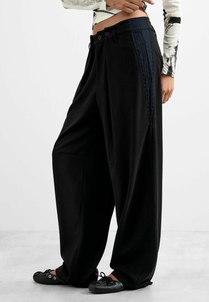 Weite schwarze Hose mit marineblauen Nadelstreifen an den Seiten, kombiniert mit schwarzen Ballerinas und einem kurz geschnittenen Langarmtop mit abstraktem Muster.