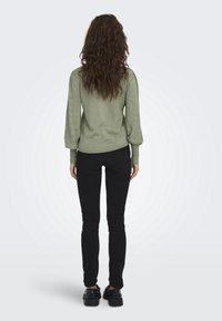 Vihreä neulepusero puffihihnoilla ja reikänauhakuosilla, yhdistettynä mustiin slim fit -farkkuihin ja mustiin kenkiin, nähtynä takaa.
