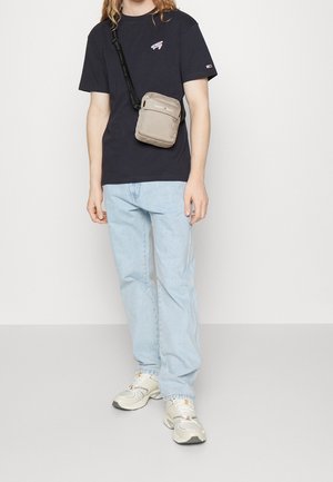 Person in einem schwarzen Tommy Hilfiger T-Shirt, hellblauen Jeans, weißen Sneakers und einer beigefarbenen Tommy Jeans Umhängetasche vor einem schlichten Hintergrund.