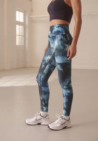Leggings de cintura alta con un patrón de acuarela azul y detalles en negro. Las mallas están hechas de un tejido elástico y texturizado. Unas zapatillas deportivas blancas completan el conjunto.