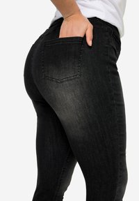 Jeans de mezclilla negra con un diseño ajustado; cuenta con un bolsillo en la parte posterior, textura suave y un ligero degradado de color de oscuro a claro.