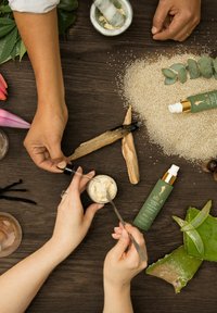 Une bouteille en verre vert avec un capuchon doré et une pompe blanche, un pot en verre de crème, des feuilles d'aloe, des morceaux de bois et des grains texturés sur une surface en bois.