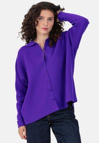Pull surdimensionné en tricot violet avec col, fermeture boutonnée sur le devant et manches longues, présentant une coupe ample et un tissu texturé.