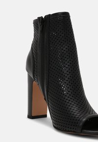 Bottine en cuir noir perforé avec bout ouvert, fermeture éclair latérale et talon haut bloc.
