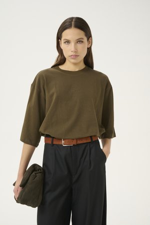 Brun oversized T-skjorte med rund hals, som har en subtil logo på brystet, kombinert med svarte bukser og et brunt belte.