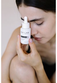 Serum Velvet Boost IDOLAB w mrożonej szklanej butelce, białej z pipetą, z czarnym napisem. Modelka trzyma go blisko twarzy.