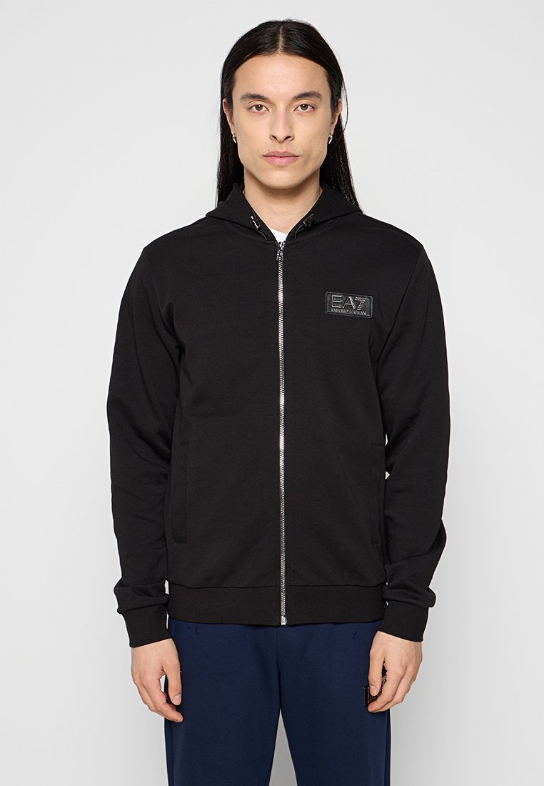 Schwarze Kapuzenjacke mit einem Logo-Patch auf der Vorderseite, mit Kapuze, Fronttaschen und einer glatten Textur. Kombiniert mit blauen Hosen.