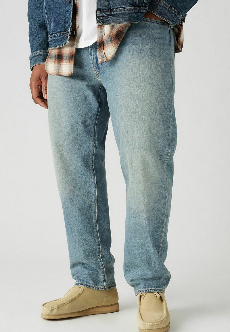 Jean en denim bleu clair avec une coupe décontractée, présentant des décolorations subtiles et des détails de couture, associés à des chaussures à enfiler en suède beige.