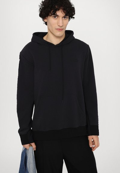 Musthave mustriibul kapuutsiga sall, millel on tasku, tekstuuriga viimistlus, lõdva lõike ja ribakäistega. On seljas mustad püksid ja käes denim-jakk.