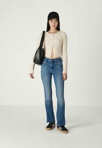 Giovane donna che indossa un berretto nero, un cardigan crema, jeans blu, sneakers nere e tiene in mano una borsa a tracolla nera, mentre si trova davanti a uno sfondo uniforme.