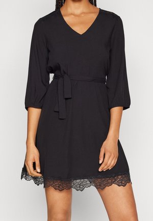 Femme portant une robe noire à col en V avec manches 3/4, ceinture à la taille et bordure en dentelle à l'ourlet, debout devant un fond uni.