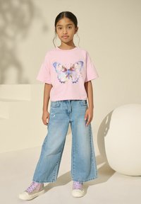 Camiseta de manga corta rosa con gráfico de mariposa, combinada con pantalones vaqueros de pierna ancha en un tono azul claro y zapatillas plateadas brillantes. Tejido suave, ajuste relajado.