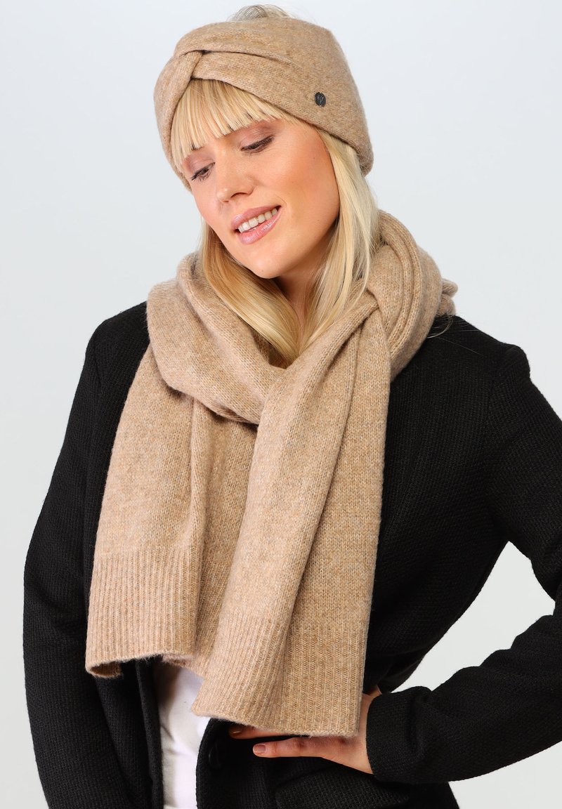 Un bandeau en tricot beige avec un design torsadé et un petit bouton d'accent, associé à une écharpe à côtes épaisses assortie, drapée sur un pull noir.