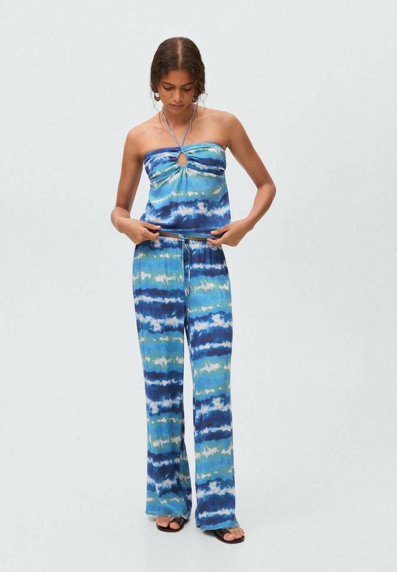 Femeie ajustând șnurul de la un top halter bleu și alb cu model tie-dye și pantaloni largi asortați, purtând sandale negre, pe un fundal simplu.