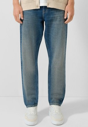 Mężczyzna ubrany w sprane niebieskie jeansy o prostych nogawkach, beżowy kardigan, białe trampki, stojący na tle jednolitego jasnego tła.