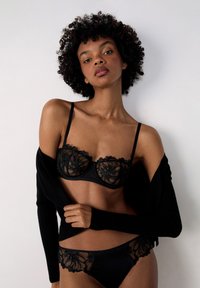 Schwarzer Spitzen-Bralette mit floralen Stickereien und Bügel, kombiniert mit passenden durchsichtigen Pantys. Weiche, strukturierte schwarze Strickjacke über einer Schulter drapiert.