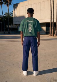 Homme debout à l'extérieur, de dos, portant un T-shirt vert avec un graphisme de vase et un texte, pantalon bleu, baskets blanches, palmiers et bâtiment à proximité.