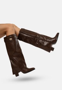 Botas de cuero marrón hasta la rodilla con tacón cuadrado y detalles de cremallera, que presentan un acento metálico en la parte superior y un acabado de textura suave.