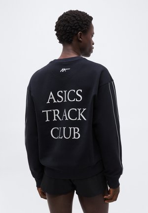 Rövid, göndör hajú személy fekete pulóvert visel, amelynek hátán az "ASICS TRACK CLUB" felirat olvasható, valamint fekete rövidnadrágot, egyszerű háttér előtt.