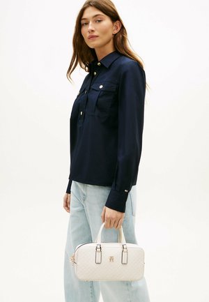 Kobieta ubrana w granatową koszulę i jasnoniebieskie dżinsy trzyma białą pikowaną torebkę ze złotymi akcentami na jednolitym tle.