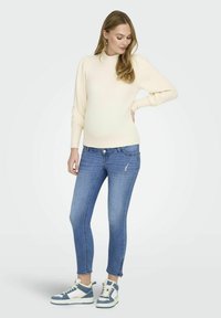 Suéter de punto acanalado crema con mangas abullonadas, combinado con jeans ajustados azules con detalles desgastados y zapatillas altas azul claro.