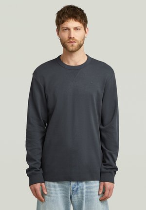 Donkergrijze trui met lange mouwen en een ronde hals, gemaakt van zachte stof met subtiele stikdetails. Gecombineerd met lichtblauwe spijkerbroek.