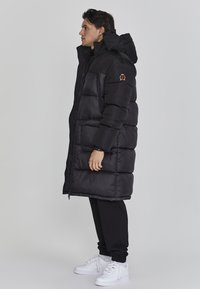Schwarze Pufferjacke mit hohem Kragen und Logopatch, mit einem gesteppten Design, durchgehendem Reißverschluss und elastischen Bündchen. Getragen über schwarzen Hosen und weißen Sneakers.